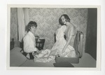 A woman holding a dress exclaiming something to a distracted cheerleader in a bedroom by Lonnie W. Fleming Sr.