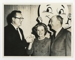 Two men and a woman in front of a smiley cartoon poster board by Lonnie W. Fleming Sr.